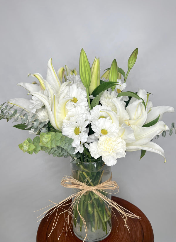 a beautiful arrangement of white daisies, white oriental lilies, cushion poms, carnations and baby blue eucalyptus all tied and in a clear cylinder vase.